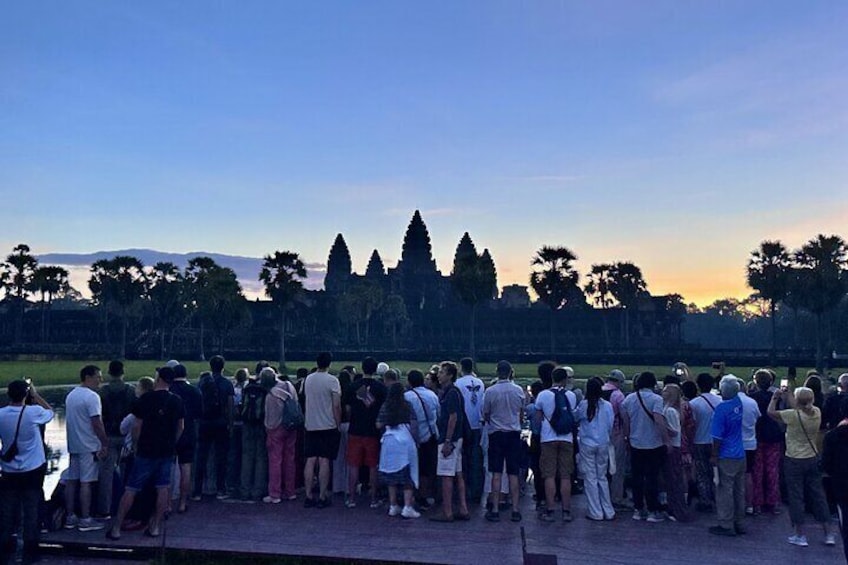 A group of people standing in a line, facing a distant view of a temple complex with tall trees and a clear sky. 1