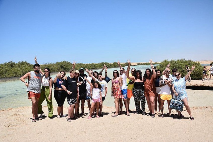 Een groep mensen poseert op een zandstrand, met water en begroeiing op de achtergrond. 3