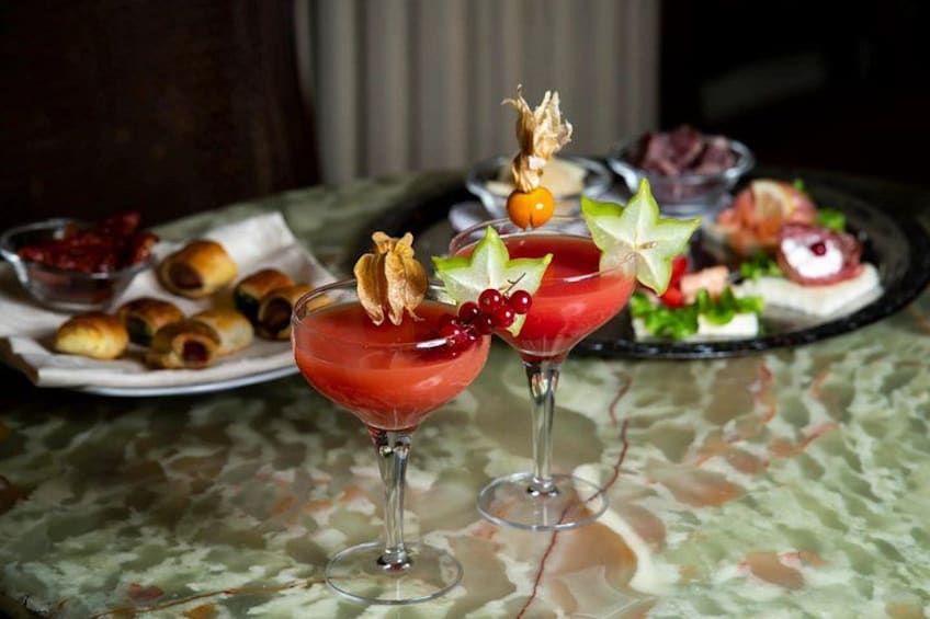 Two martini glasses with red cocktails, garnished with fruit and edible flowers, on a table with a patterned tablecloth. 2