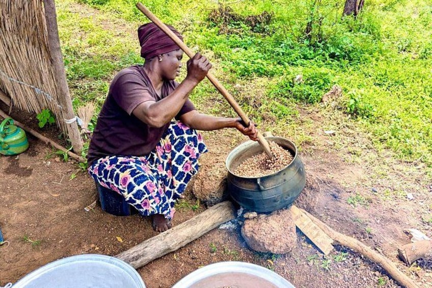 A person sitting outdoors, stirring contents in a large pot with a long stick. 1