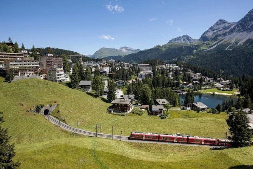 A red train on tracks, a town with buildings, a lake, and mountains in the background. 4