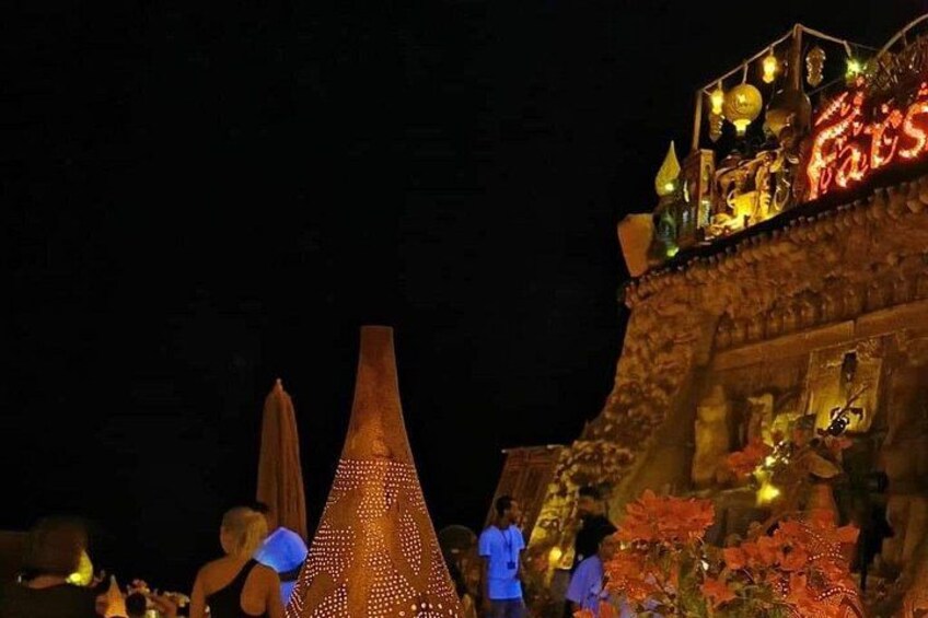 A night scene with illuminated structures, including a large cone-shaped object and a building with a lit sign. 5