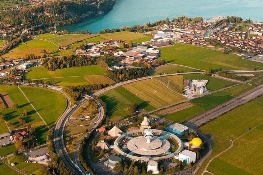 An aerial view of a town with a circular building complex, roads, fields, and a body of water. 3