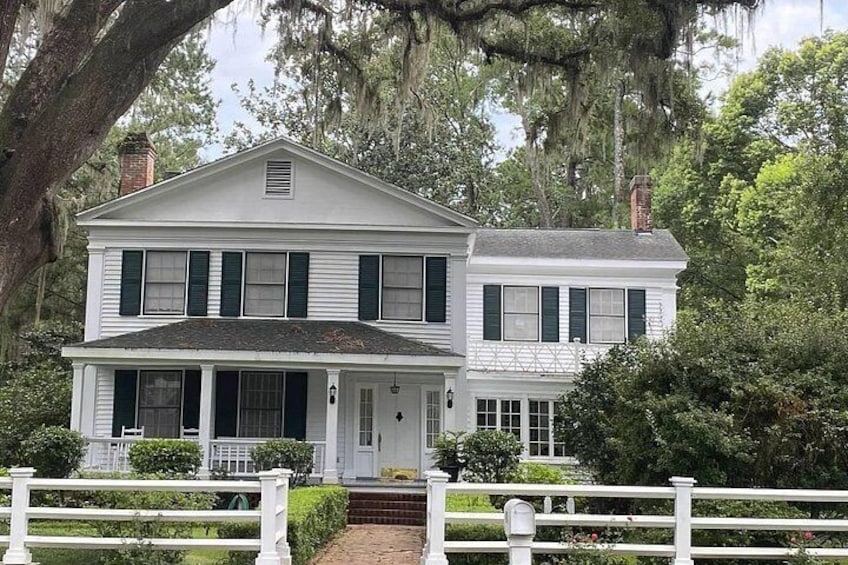 A white two-story house with green shutters, a white porch, and a white fence in front. 1