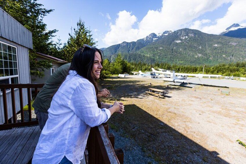 Une personne se tient sur une terrasse en bois, surplombant une zone de gravier, avec des avions et des montagnes en arrière-plan. 3