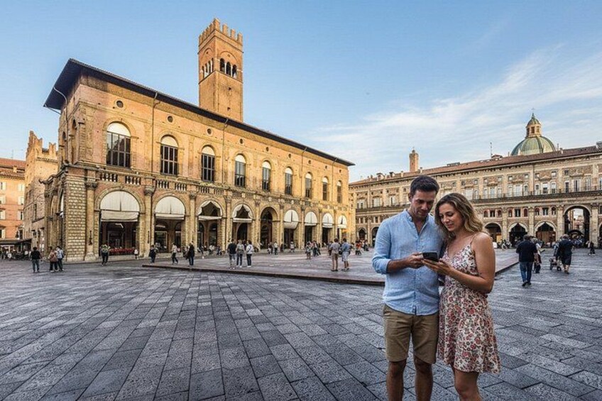 A historic building with a tower and arched windows, a paved square, and people walking. 4