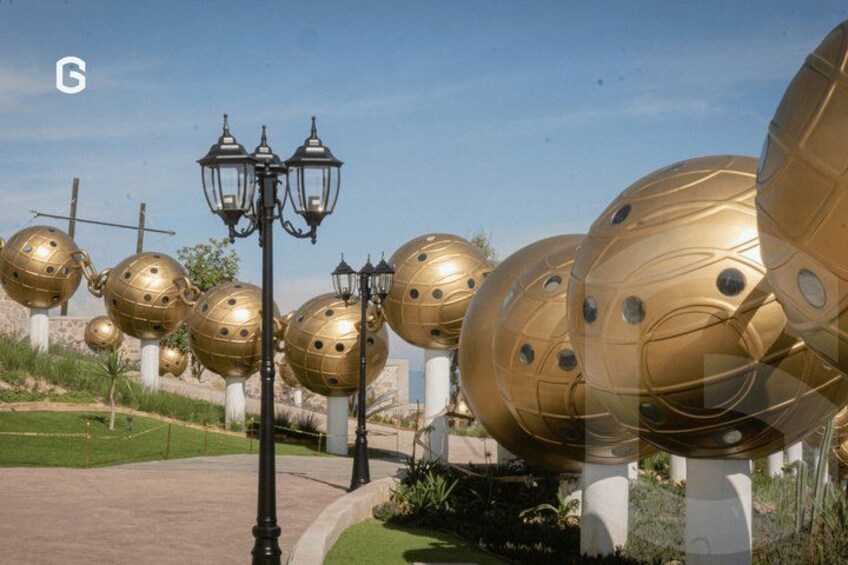 Golden spherical sculptures, a street lamp, and a cross in a landscaped area. 4