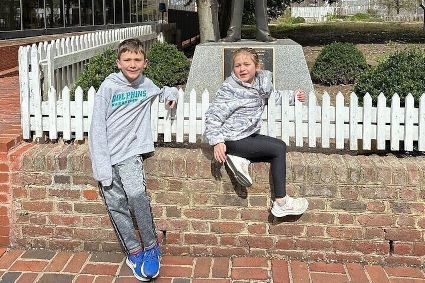 Two children standing in front of a brick wall with a white picket fence in the background. 1