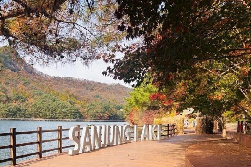 A lakeside walkway with a wooden fence, trees, and the text 'SANJUNGAI' on the ground. 2