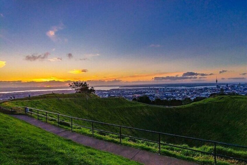 Una colina cubierta de césped con un camino y barandilla, con vistas a un paisaje urbano al atardecer. 5