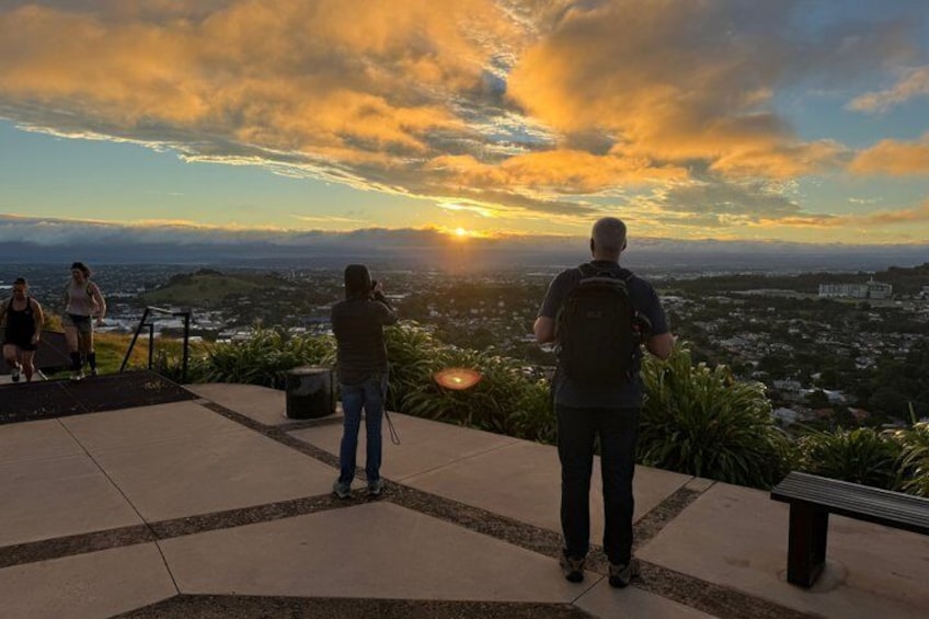 Vista del atardecer desde una terraza con gente de pie tomando fotografías. 1