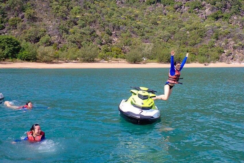 En person hopper fra en jetski ned i vandet, en anden person svømmer i nærheden, og en strand med træer i baggrunden. 2