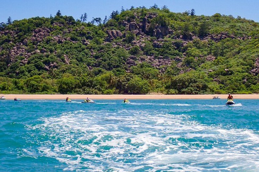 Ein Strand mit klarem türkisfarbenem Wasser, ein felsiger Hügel im Hintergrund und mehrere Menschen in kleinen Booten. 2