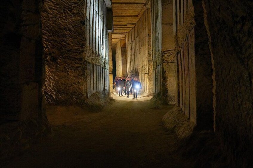 Un pasaje estrecho y tenuemente iluminado, con paredes de piedra y vigas de madera, que conduce a un grupo de personas con linternas. 1