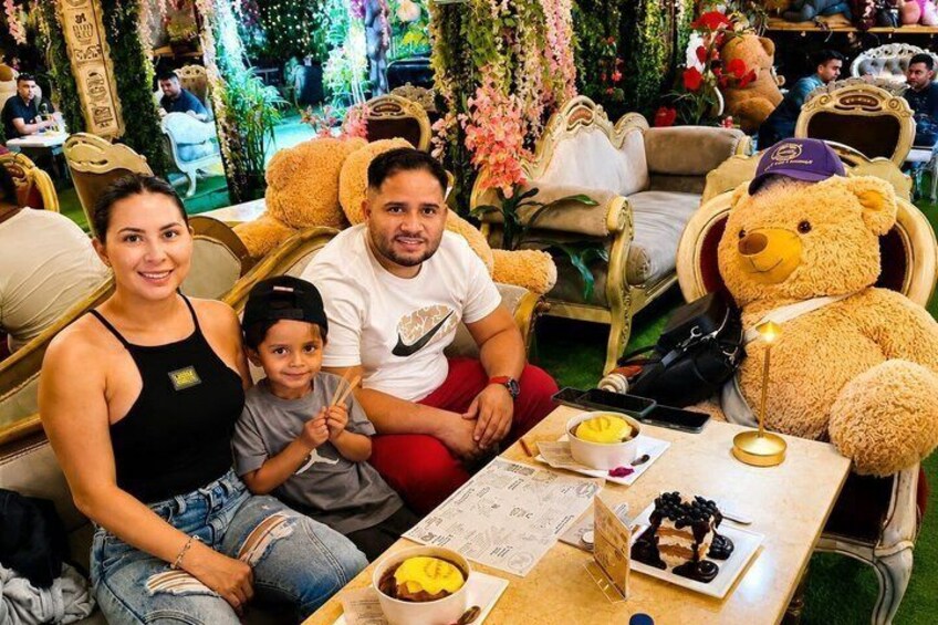 Eine Familie sitzt an einem Tisch mit einem Teddybären, umgeben von verzierten Stühlen und floralen Dekorationen. 2
