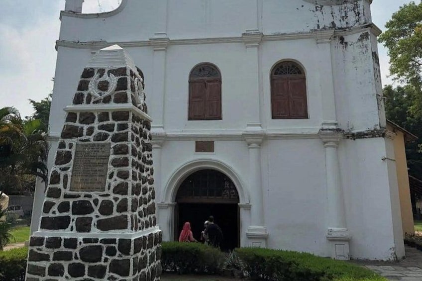 Eine weiße Kirche mit einem steinernen Denkmal davor, Holztüren und einem kleinen Rundbogenportal. 3