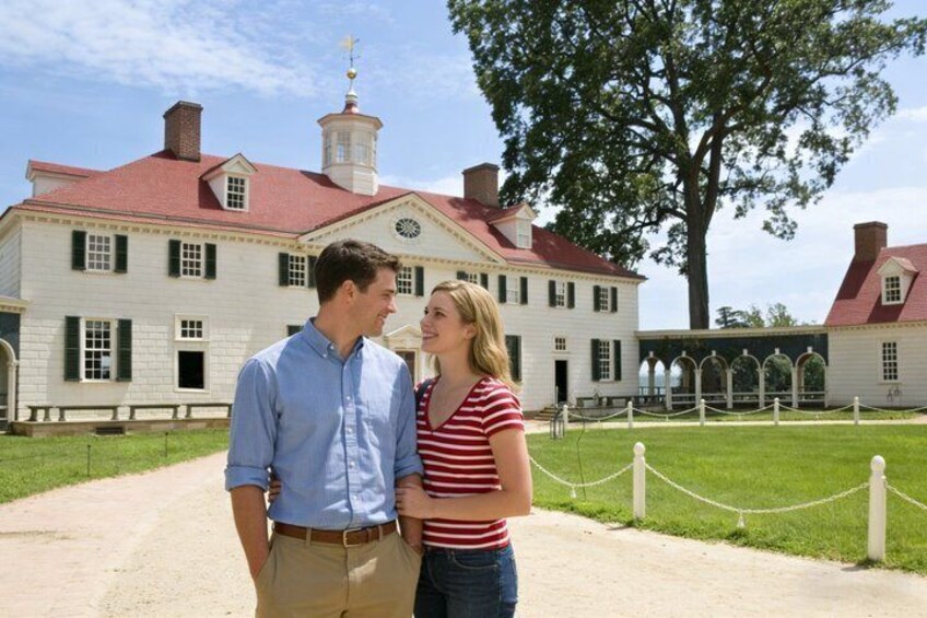 A historic building with a red roof and white walls, a couple standing in front, and a tree to the right. 2