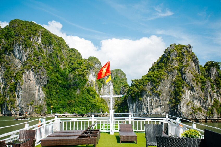 A boat deck with wicker chairs and tables, a flagpole with a flag, and a backdrop of limestone cliffs and greenery. 5