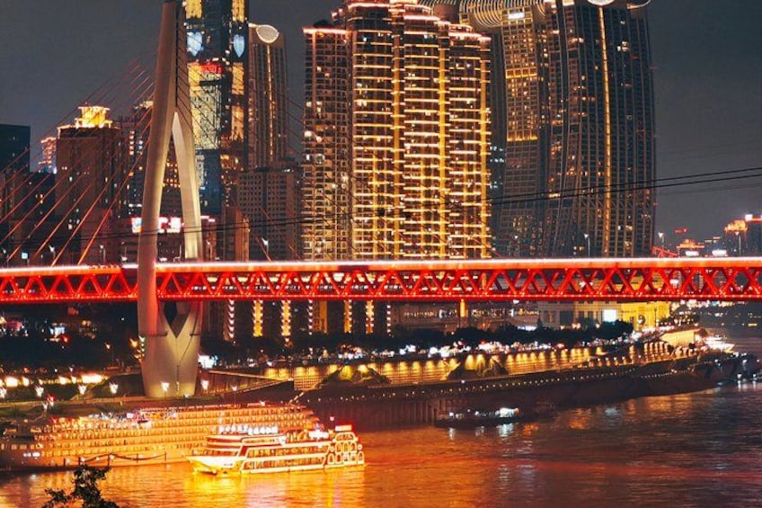 Une vue nocturne d’une ville, avec un pont rouge bien visible, des bâtiments illuminés et une rivière où naviguent des bateaux. 2