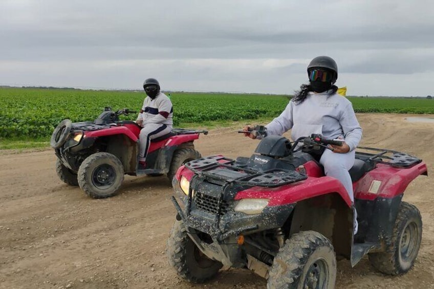 Two people on red all-terrain vehicles on a dirt road with green fields in the background. 1