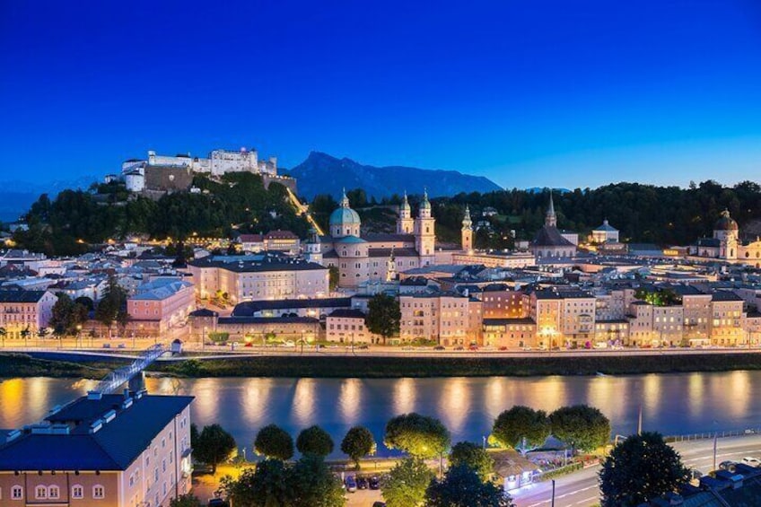 Un paisaje urbano al atardecer con edificios iluminados, un río y un castillo en una colina. 1
