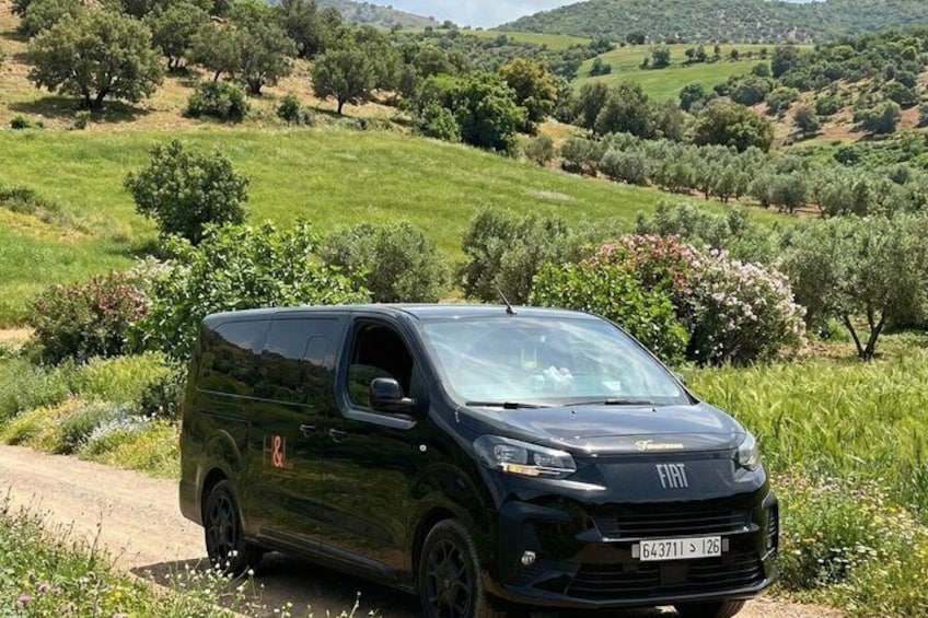 Een zwarte Fiat bestelwagen staat op een onverharde weg in een landelijk gebied met groene velden en bomen. 2