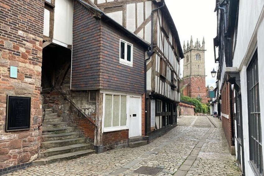 A cobblestone street lined with timber-framed buildings and a church tower in the distance. 1
