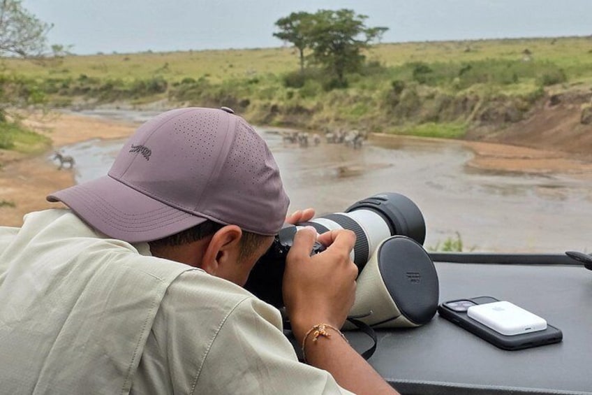 A person using a camera with a large lens, a smartphone on the table, and a natural landscape with a river and trees in the background. 4