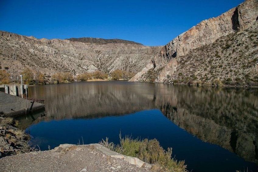 A calm lake with a rocky shoreline, a dam structure, and a clear blue sky. 3