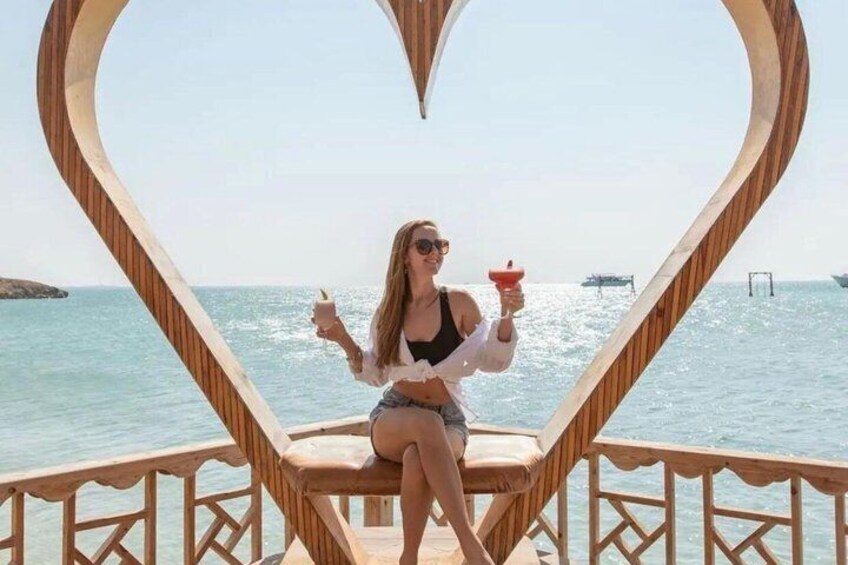 A person sitting on a wooden deck chair with a heart-shaped frame, holding a drink, with a body of water and boats in the background. 2