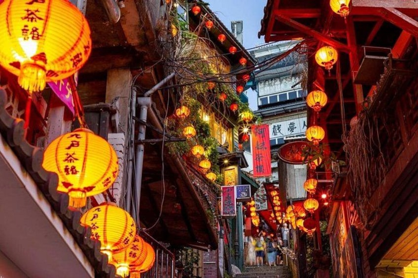 A narrow street with traditional buildings, hanging lanterns, and red banners. 3