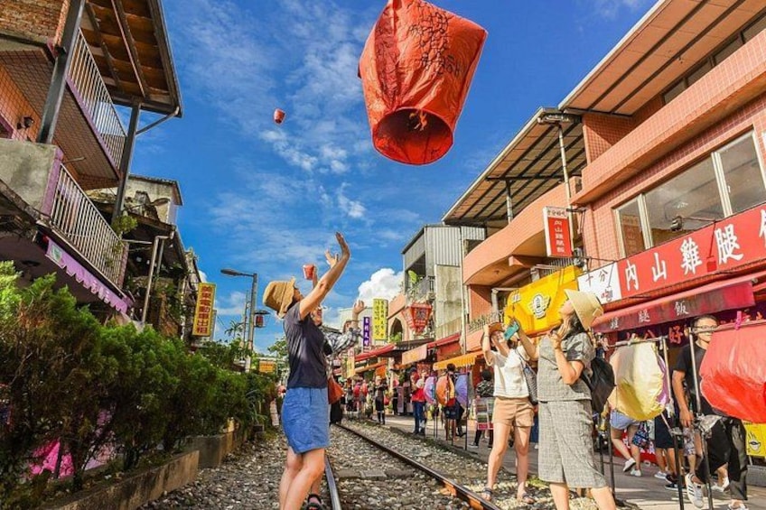 A street with people releasing red lanterns, a train track, and buildings with signs. 1