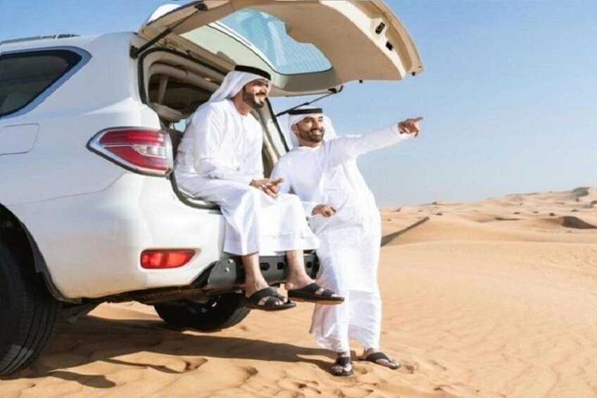 Zwei Männer in traditioneller Kleidung sitzen auf der Heckklappe eines weißen SUVs in einer Wüste. 5