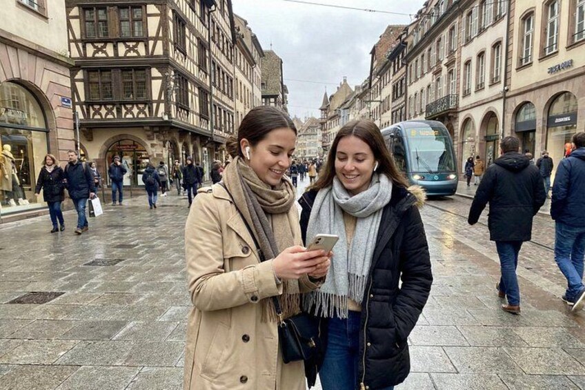 Deux personnes se tiennent debout sur une rue pavée et mouillée, avec des bâtiments historiques et un tramway en arrière-plan. 4