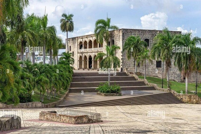 A historic building with arched windows and a staircase leading up to it, surrounded by palm trees and a paved area. 2