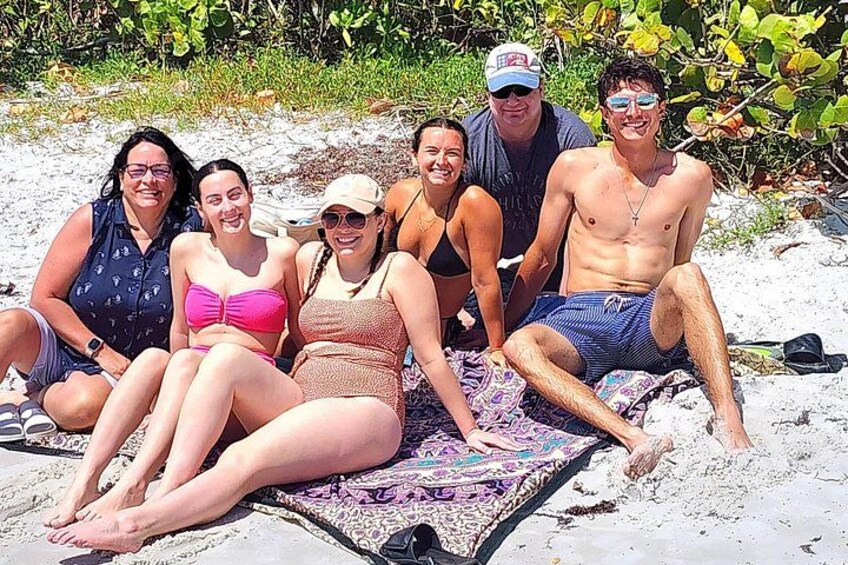 A group of people sitting on a blanket on a sandy beach with vegetation in the background. 4
