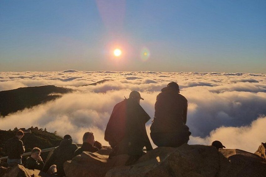 People sitting on a mountain ridge overlooking a sea of clouds with the sun setting in the background. 4
