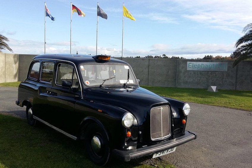 A vintage black car parked on a paved area with grass on the sides. There are flags in the background and a sign that reads 'Elephant.' 2