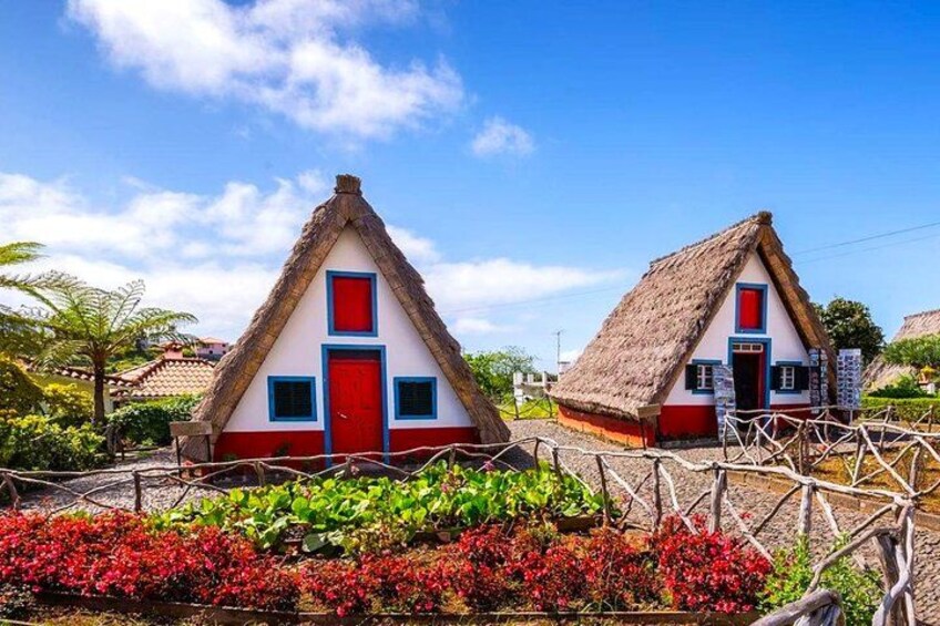 Dos casas con techos de paja, puertas rojas y paredes blancas, rodeadas de flores coloridas bajo un cielo azul despejado. 1