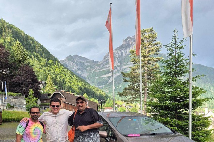 Three people standing in front of a car with flags, a mountain, and a lake in the background. 2