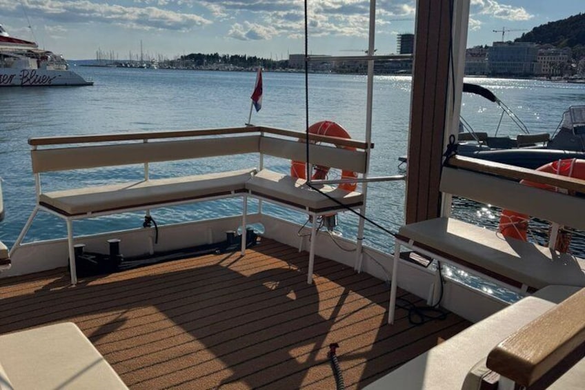 Un pont de bateau équipé de sièges et de bouées de sauvetage, un drapeau, et une vue sur l’eau avec d’autres bateaux et une côte lointaine. 4
