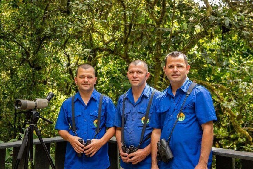 Three people in blue shirts standing in a forested area with binoculars and a camera on a tripod. 2