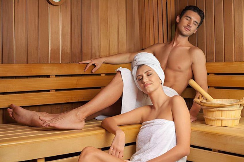 Zwei Personen in einer Sauna mit Holzbänken und einem Holzeimer. 3