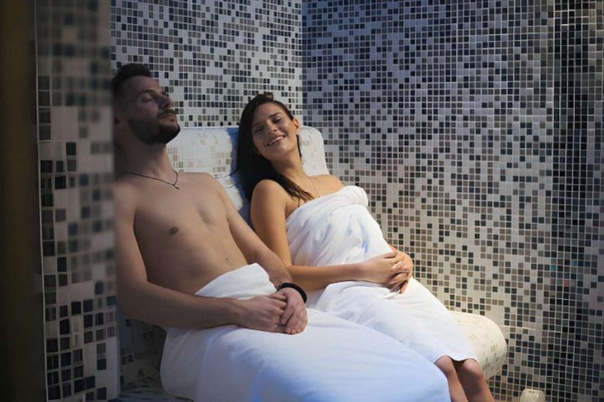 Ein Paar in einem mit Fliesen ausgelegten Raum, möglicherweise ein Spa- oder Badebereich, mit einer Wand aus Mosaik. 2