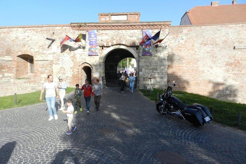 A cobblestone pathway leading to a historic stone gate with flags, a motorcycle parked nearby, and people walking. 2