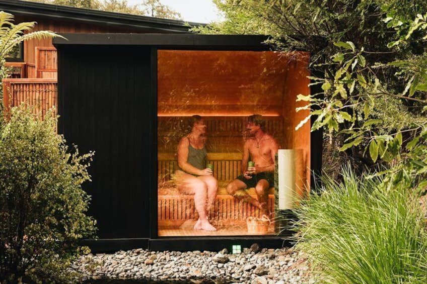 A wooden sauna structure with two people inside, surrounded by greenery. 4