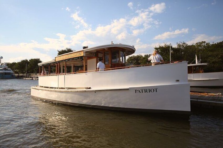Een witte boot genaamd 'PATRIOT' afgemeerd in een jachthaven, met een overdekt dek en een persoon op het voordek. 1