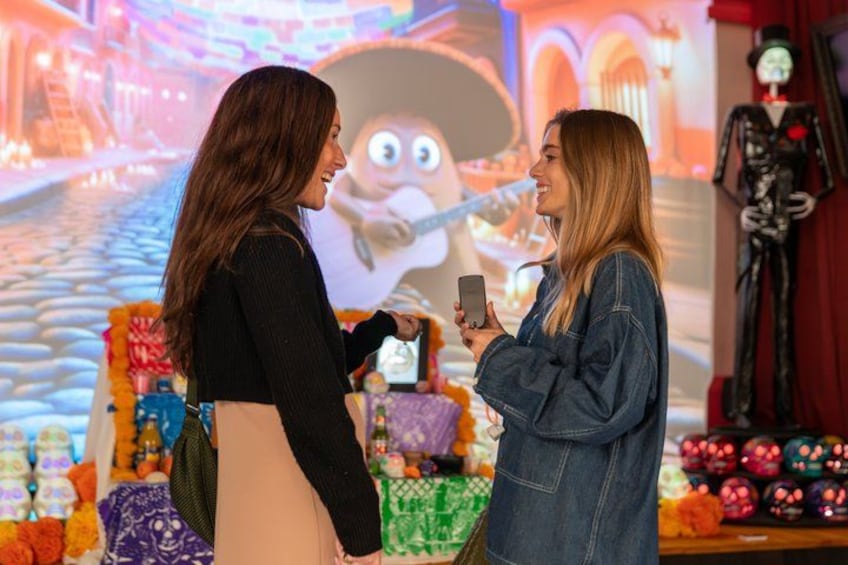 Dos personas en un ambiente festivo, con un personaje animado de gran tamaño y una figura de esqueleto al fondo. 3