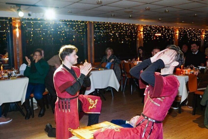 Two individuals in red costumes with gold accents, possibly performing or engaging in a ceremonial act, in a dimly lit indoor setting with tables and chairs. 3