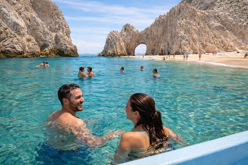 A couple in a clear blue pool with rocky cliffs and a beach in the background. 1